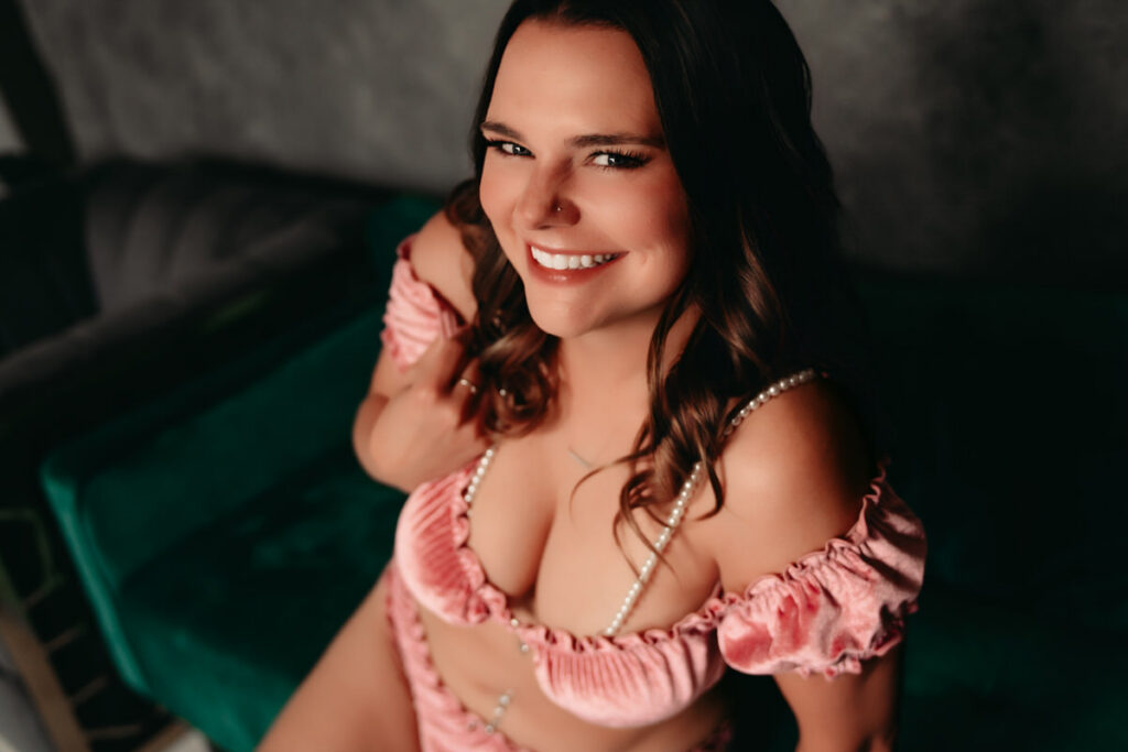 Woman smiling and sitting on a green sofa with pink two piece lingerie with pearls.