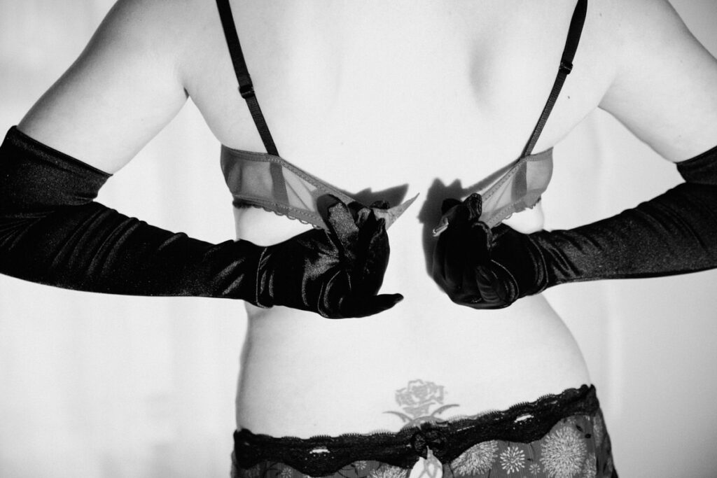 black and white image of a woman with a tattoo on her lower back and she is undoing her bra with gloves on